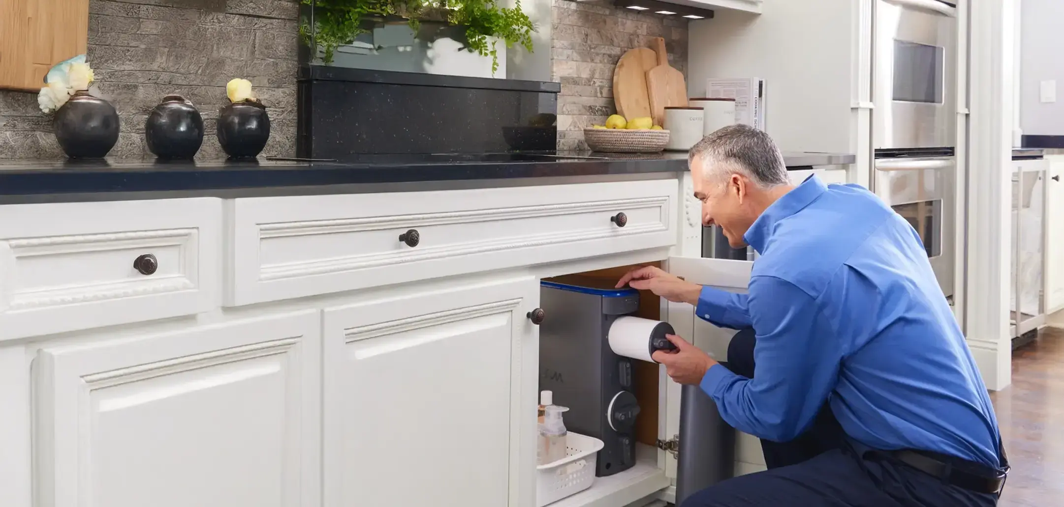 A man in a blue button-down shirt kneeling in a modern kitchen, replacing a white cylindrical filter in a grey under-sink water filtration system housed within a white cabinet.