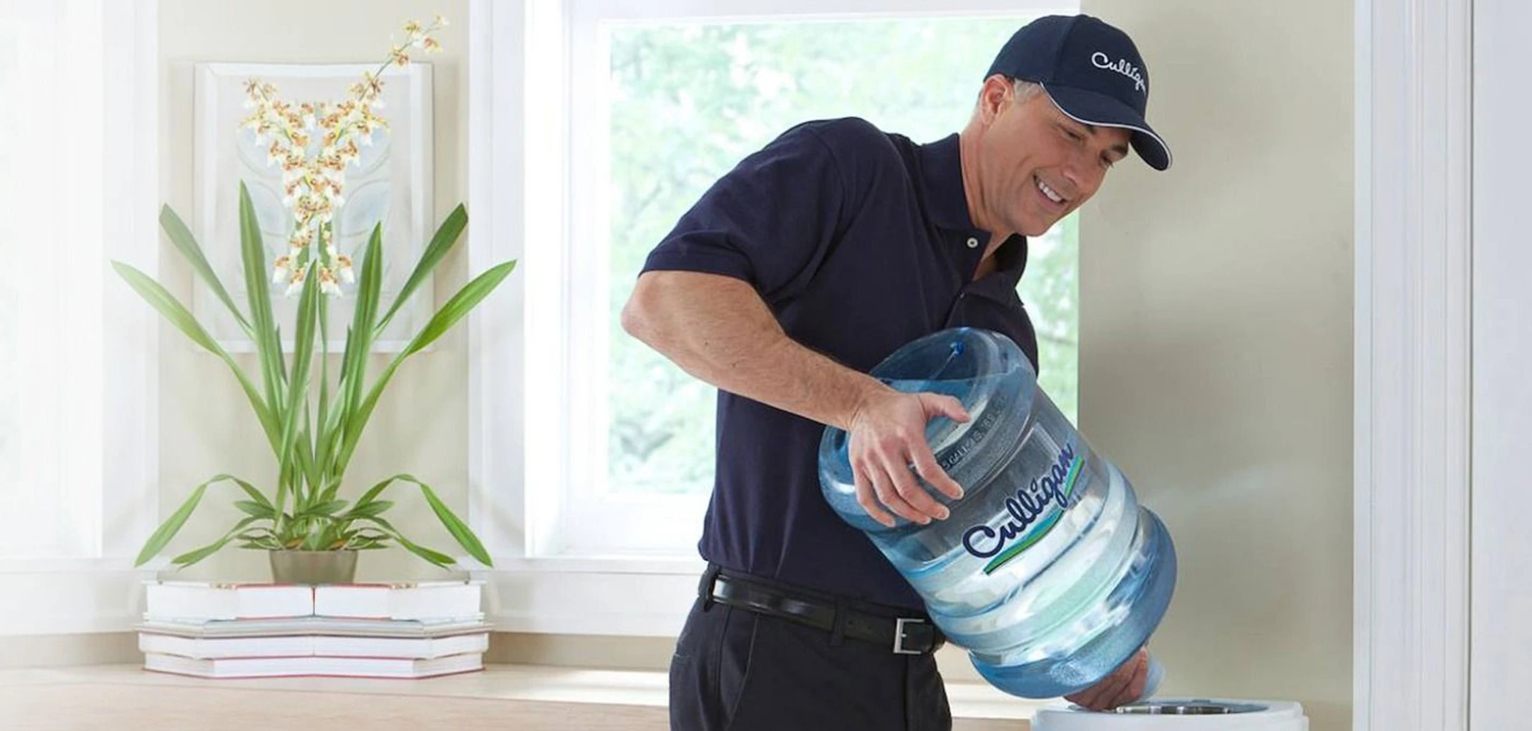 A smiling Culligan service professional in a blue uniform and cap placing a large 5-gallon water bottle onto a white top-loading water cooler in a bright, modern room with a window and a potted orchid.