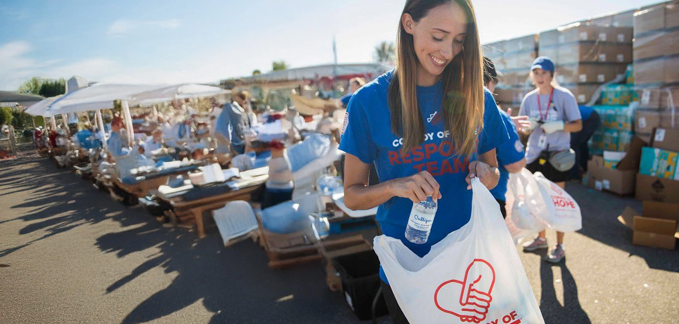 A smiling female volunteer in a blue 'Response Team' shirt places a Culligan water bottle into a white bag with a red heart logo. In the background, other volunteers and people are gathered around tables under canopies, with stacks of relief supplies visible further back under a bright sky.