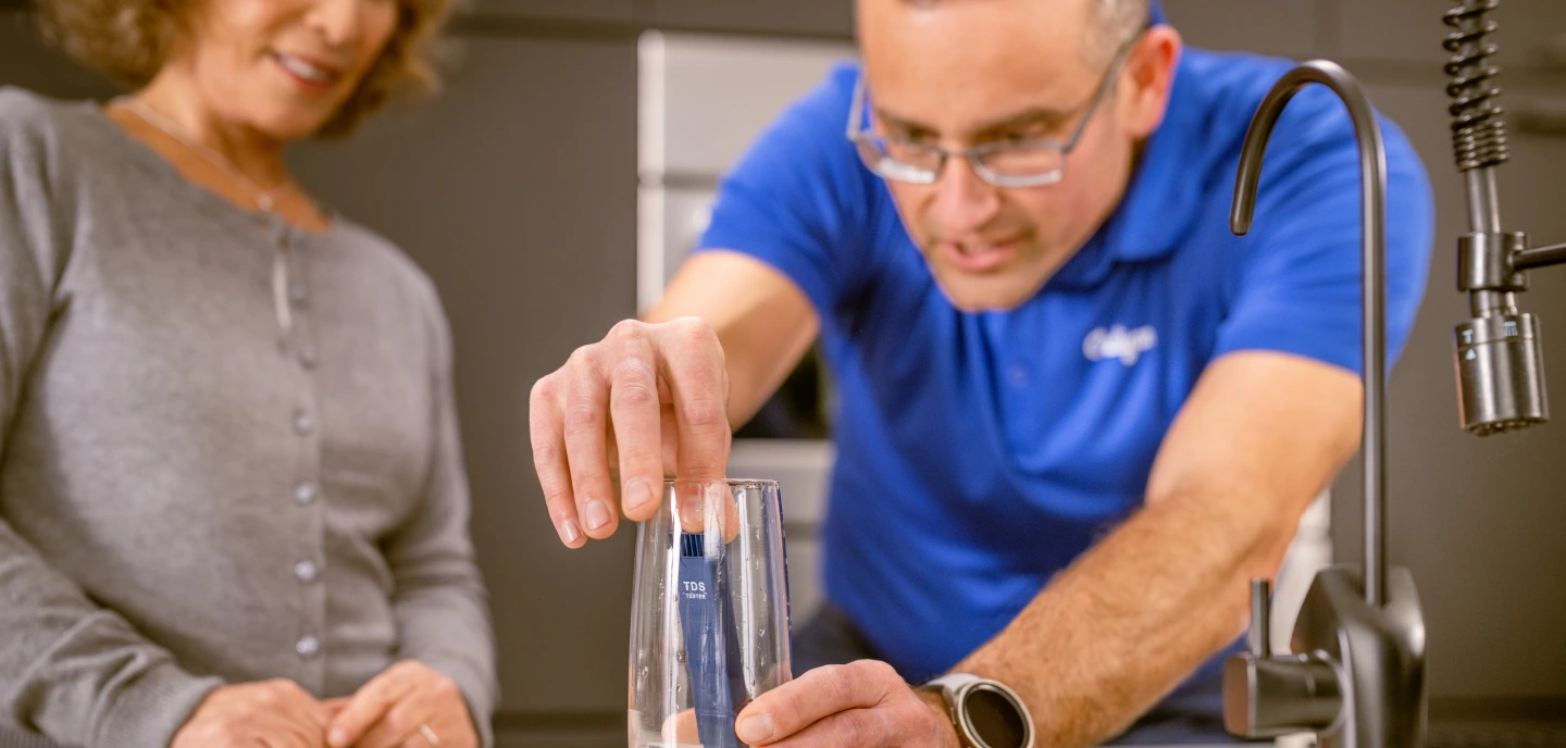 A man in a blue shirt uses a blue TDS tester in a glass of water, while a woman watches. They are in a modern kitchen.