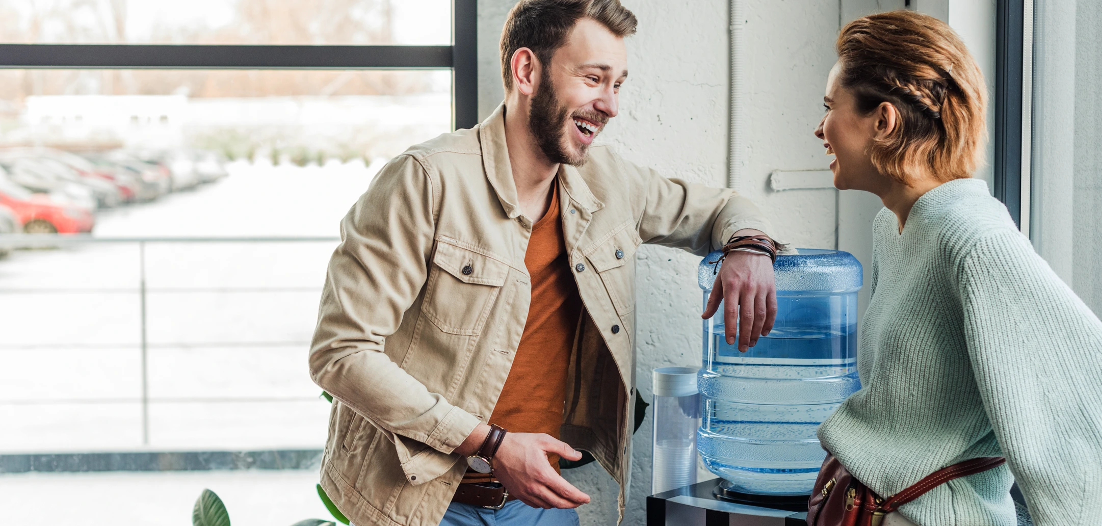 Two smiling people, a man and a woman, laughing while talking by a water cooler in an office with a snowy outdoor view.