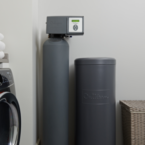 A Culligan water softener system, consisting of two dark gray tanks, installed in a clean utility room next to a washing machine and a stack of white towels.