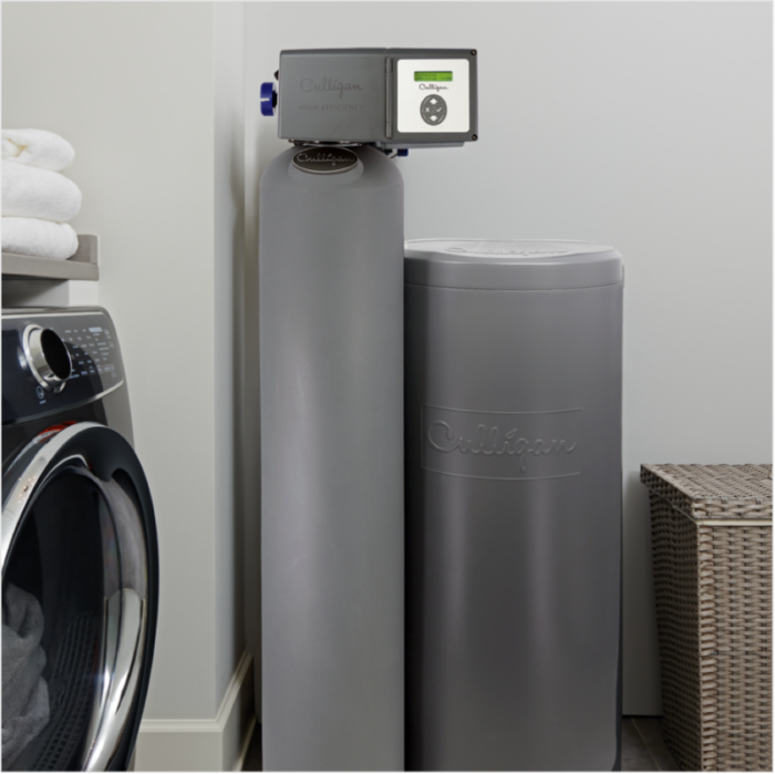 A Culligan water softener system, consisting of a main tank with a digital controller and a brine tank, stands in a laundry room next to a washing machine and folded towels.
