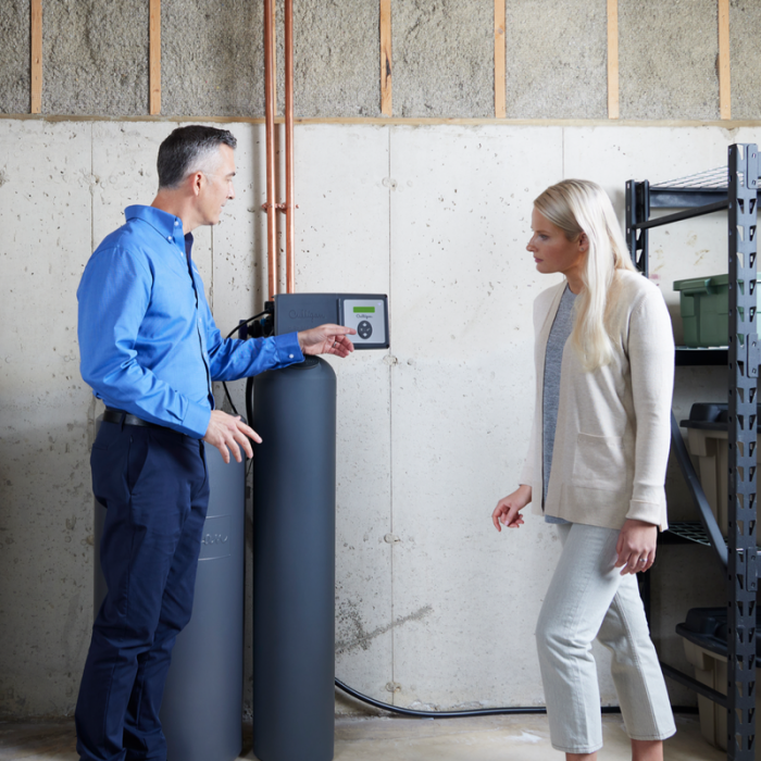 A man in a blue shirt explains a water treatment system to a woman in a beige cardigan in a basement.