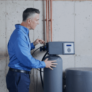 A man in a blue shirt adjusts the control panel of a gray Culligan water softener unit in a utility room with concrete walls and copper pipes.