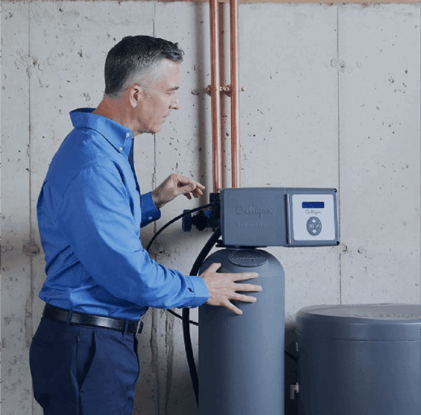 A man in a blue shirt adjusts the control panel of a gray Culligan water softener unit in a utility room with concrete walls and copper pipes.