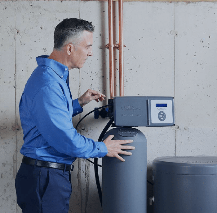 A man in a blue shirt adjusts the control panel of a gray Culligan water softener unit in a utility room with concrete walls and copper pipes.
