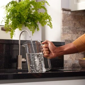 A hand holds a glass pitcher under a modern kitchen faucet, filling it with water. A green fern plant and stone backsplash are in the background.