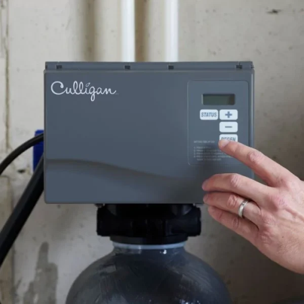 A hand presses the 'REGEN' button on a gray Culligan water treatment system control panel, which sits atop a large gray tank.