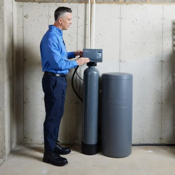 A man in a blue shirt and dark pants adjusting the control panel of a gray Culligan water softener system in a concrete basement.
