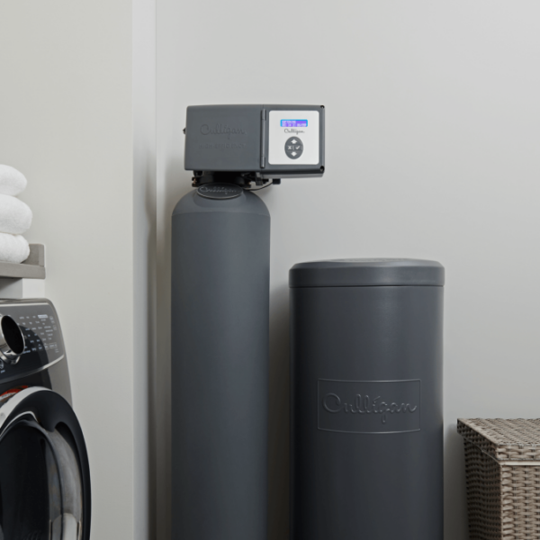 A dark gray Culligan water softener and matching brine tank are installed in a laundry room, next to a washing machine and a stack of white towels.