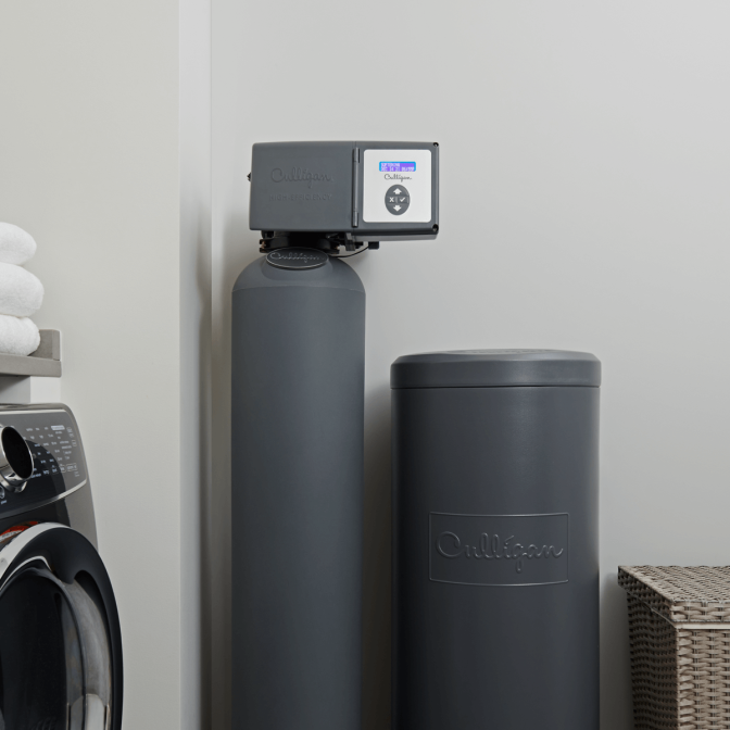 A dark gray Culligan water softener and matching brine tank are installed in a laundry room, next to a washing machine and a stack of white towels.