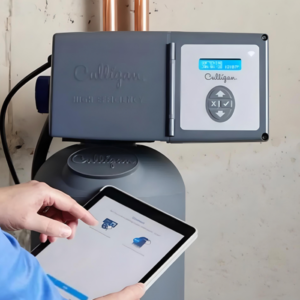A person's hands using a tablet to control a gray Culligan High-Efficiency water treatment system, which has a digital display and copper pipes.