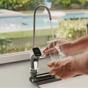 A person's hands fill a clear glass with water from a modern chrome Culligan faucet, which has a digital display showing a green and blue graphic. A swimming pool and greenery are visible through a window in the background.