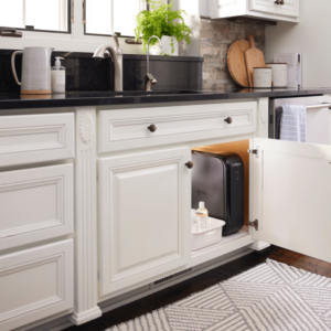 A modern kitchen with white cabinets, black countertops, and a black Culligan water filtration system hidden inside an open under-sink cabinet.